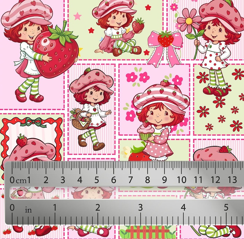 May include: A fabric print featuring Strawberry Shortcake characters in various poses with strawberries, flowers, and cupcakes, shown with a ruler measuring up to 5 inches and 13 cm to indicate the scale of the repeating pattern on the material.