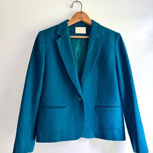 May include: A teal blazer featuring a single-button closure, notched lapels, and two horizontal welt pockets. The garment is lined with a matching teal fabric and includes a white interior brand label at the neckline, displayed on a wooden hanger.