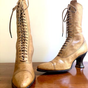May include: A pair of vintage tan leather boots with a lace-up design. The boots have pointed toes, high heels, and are displayed on a wooden surface. The boots are a light brown color and appear to be in good condition.