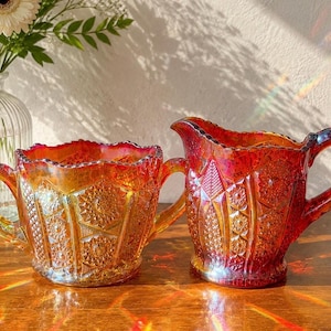 May include: A pair of iridescent marigold carnival glass pieces consisting of a two-handled sugar bowl and a matching creamer pitcher. Both items feature a pressed geometric starburst pattern with a scalloped rim and a reflective orange finish.