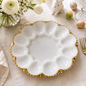 May include: A round white ceramic egg serving platter featuring twelve recessed oval indentations for holding eggs. The plate has a scalloped edge finished with a metallic gold trim that highlights the curved design of the serving dish.