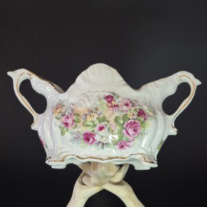 May include: A white porcelain bowl with two ornate handles and gold-trimmed edges. The center features a detailed floral pattern with pink roses, green leaves, and small yellow flowers. The bowl has a scalloped rim and a curved, decorative shape.