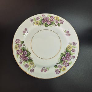 May include: A round white ceramic plate featuring a decorative border of purple and yellow flowers with green leaves. The center of the plate is plain white and is encircled by a thin gold rim, with an additional gold trim along the outer edge.