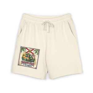Può includere: Pantaloncini color bianco sporco con elastico in vita e coulisse. Una grafica quadrata in basso a sinistra raffigura un alligatore verde con occhiali da sole che tiene una piccola bandiera americana, con la bandiera della Florida e la scritta The Freedom State of Florida.