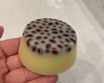 Puede incluir: Un pequeño postre redondo con una base amarilla clara y una parte superior blanca salpicada de pequeños puntos rojos oscuros. El postre se sostiene en una mano, mostrando su tamaño y forma. La apariencia general sugiere un dulce.