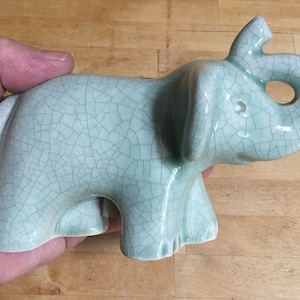 May include: A light green ceramic elephant figurine with a crackled glaze finish. The elephant's trunk is curled up and the figurine is standing on a wooden surface.