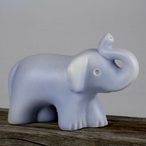 May include: A light blue ceramic elephant figurine. The elephant has a trunk raised in the air, large ears, and a small tail. The figurine is sitting on a weathered wooden surface. The elephant is a solid color with a matte finish.