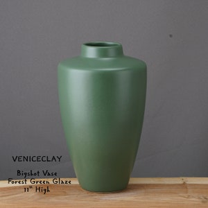 May include: A forest green ceramic vase with a simple, elegant design. It is 11 inches tall and is called the Bigshot Vase.