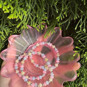 Könnte beinhalten: Drei pastellfarbene Armbänder mit silberfarbenen Akzenten liegen in einer rosa, blütenförmigen Schale. Die Armbänder haben rosa, gelbe und blaue Perlen. Die Schale steht vor einem Hintergrund aus grünem Laub.