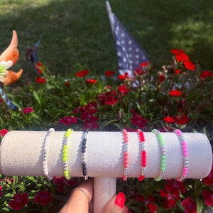 May include: Seven beaded bracelets displayed on a beige T-bar stand. The bracelets feature alternating patterns of small metallic silver beads and colorful round beads in white, neon yellow, black, coral, red, neon green, and bright pink.