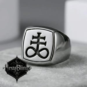 May include: A silver-toned ring with a square face featuring a black symbol. The ring has a polished finish and a thick band. The brand name "Hexy Blings" is visible in a black and white logo in the lower left corner.