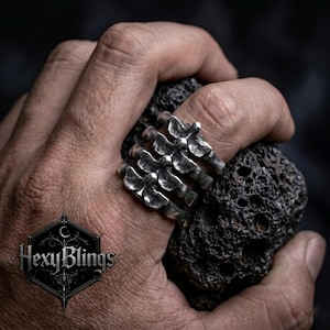 May include: A silver-colored ring shaped like a skeletal hand, with articulated finger bones. The ring is worn on a finger and held against a dark, textured stone. The brand logo "HexyBlings" is visible in the lower left corner.