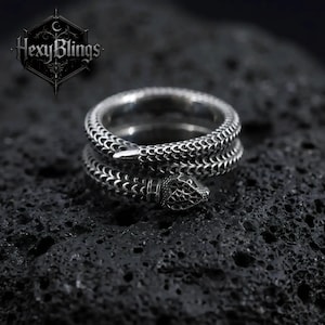 May include: A silver snake ring, coiled and detailed with scale patterns. The snake's head is raised, and the ring sits on a dark, textured surface. The brand name "HexyBlings" is visible in the upper left corner of the image.