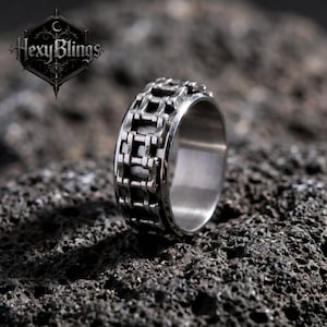 May include: A silver-colored ring with a chain design. The ring features a detailed pattern of interconnected chain links around the band. The ring is set against a dark, textured background. The brand name "Hexy Blings" is visible in the upper left corner.
