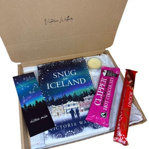 May include: A gift box containing a book titled "Snug in Iceland", a bookmark with a starry sky design, a Clipper hot chocolate packet, a Lindor chocolate bar, and a small candle. The box is brown with the name "Victoria Walker" printed on the inside.