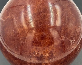 May include: A polished, spherical stone in a deep reddish-brown hue with visible internal veining and natural fractures. The surface is smooth and reflective, showing light highlights. The sphere rests upon a small, carved wooden stand base.