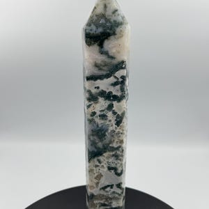 May include: A tall, rectangular moss agate crystal point. The crystal features a mix of white, gray, and green colors. A small, rounded, white stone is affixed to the top of the crystal. The crystal is standing on a black surface.