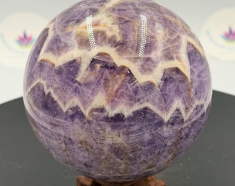 May include: A polished spherical amethyst crystal featuring distinct chevron patterns of deep purple and white quartz. The stone displays a smooth, rounded surface with natural mineral inclusions and sits atop a small, carved wooden display stand.