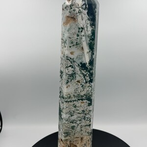 May include: A tall, polished stone obelisk with a pointed top. The stone features a mix of green, white, and tan colors, with a textured surface. The obelisk is standing on a black circular base. The overall theme is crystals and minerals.