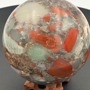 May include: A polished gemstone sphere with a mix of red, gray, and white colors. The sphere is supported by a small, carved wooden stand. The stone has a marbled appearance with various patterns and textures.