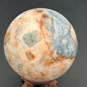 May include: A polished stone sphere with a mix of colors, including beige, gray, and rust. The sphere is displayed on a small, carved wooden stand. The stone's surface has a marbled appearance.