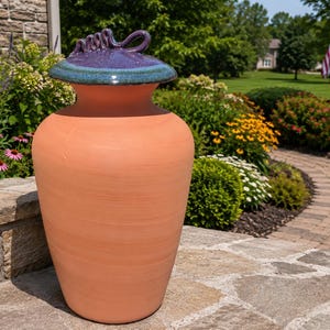May include: A terracotta-colored ceramic jar with a wide, rounded body and a narrow neck. The jar features a glossy, dark purple and teal glazed lid with a decorative, twisted handle on top, contrasting with the matte finish of the base.