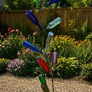 May include: A metal sculpture of a tree with colorful glass bottles as leaves. The bottles are various colors, including blue, green, and red. The sculpture is in a garden setting with flowers and a wooden fence.