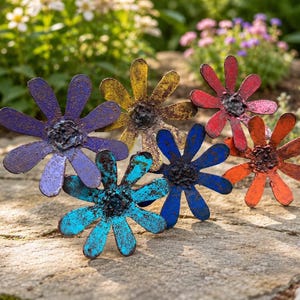 May include: A collection of six metal flower sculptures in various colors, including purple, yellow, red, orange, blue, and turquoise. The flowers have a rustic, weathered appearance and are arranged on a stone surface, with a blurred garden background.
