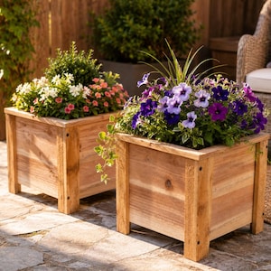 May include: Two wooden rectangular planters filled with colorful flowers. One planter has white and pink flowers, and the other has purple and blue flowers. The planters are made of light brown wood and are placed on a stone patio.