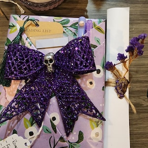 May include: A gift wrapped in light purple paper with a green leaf pattern, featuring a large glittery purple bow with a silver skull center. A white scroll tied with twine and dried purple flowers sits beside it, along with a reading list tag.