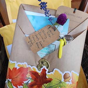 May include: A brown paper package sealed with a wax stamp and decorated with a colorful autumn leaf and acorn sticker. It contains a reading list card, a tea bag, a yellow pen, a diamond-shaped charm, and a tag with handwritten book notes.