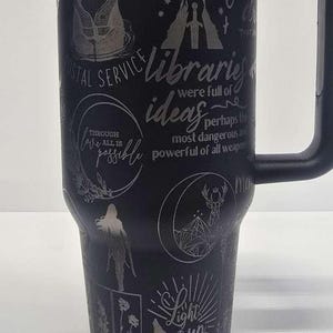 May include: Black travel mug with a handle, featuring etched designs and text including "Crescent City", a deer, and the phrase "Light it up". The mug has a black lid and a silver interior.