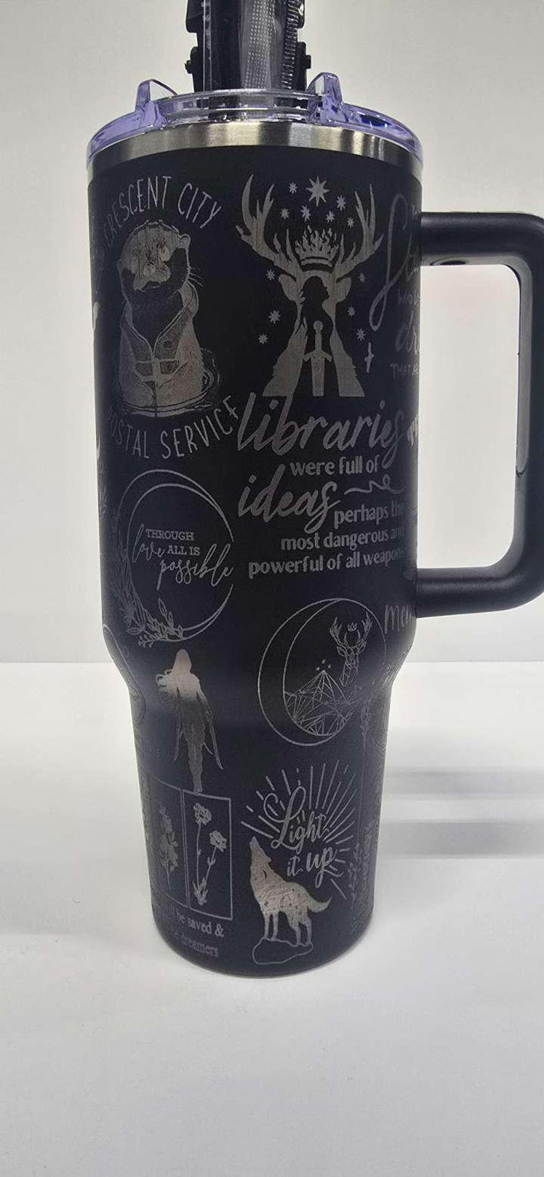 May include: Black travel mug with a handle, featuring etched designs and text including "Crescent City", a deer, and the phrase "Light it up". The mug has a black lid and a silver interior.