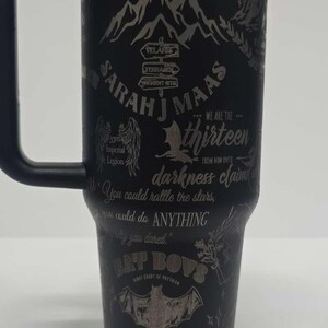 May include: Black insulated tumbler with a handle, featuring etched text and graphics. The design includes the words "THE WORLDS OF SARAH J MAAS", a mountain range, and other text and symbols. The tumbler has a black lid.