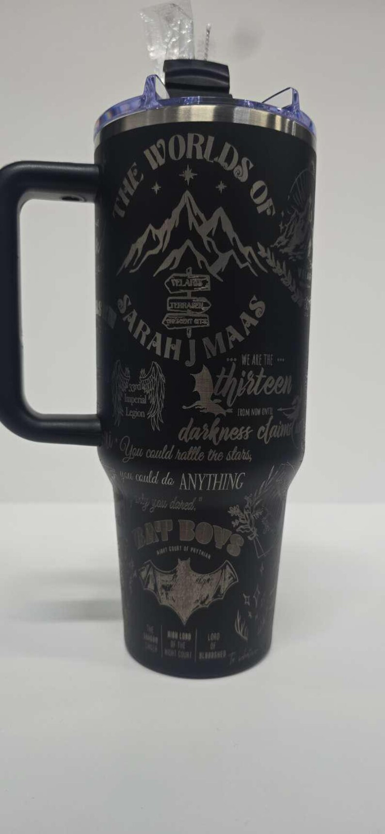 May include: Black insulated tumbler with a handle, featuring etched text and graphics. The design includes the words "THE WORLDS OF SARAH J MAAS", a mountain range, and other text and symbols. The tumbler has a black lid.