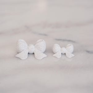 May include: Two white bows with a subtle speckled texture. The bows are shaped with a central knot and two loops on each side, with two short tails extending downward. The larger bow is positioned on the left, and the smaller bow is on the right.