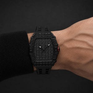 Peut inclure: Une montre-bracelet noire avec un cadran rectangulaire texturé et un bracelet noir assorti. La montre a un accent rouge sur le côté et est portée au poignet. Le cadran de la montre présente un motif quadrillé.