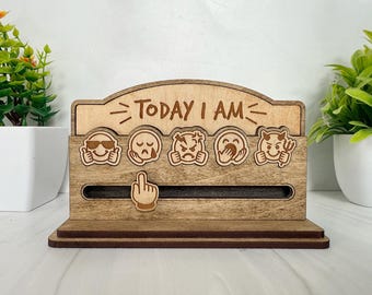May include: Wooden mood tracker with the text "TODAY I AM" and various emoji faces. A sliding pointer indicates the current mood. The emojis include sunglasses, crying, angry, covering mouth, and devil.