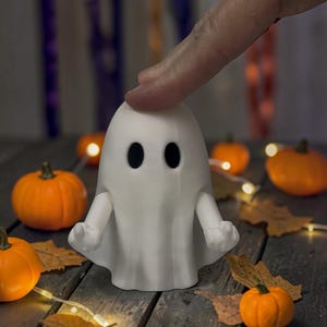 Op de afbeelding: Een wit spookvormig beeldje met twee zwarte ovale ogen en twee uitgestrekte armen ontworpen om voorwerpen vast te houden. Het spook heeft een glad, mat oppervlak en een afgeronde bovenkant, staand met een gedrapeerd uiterlijk.