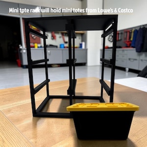 Op de afbeelding: Een zwart opbergrek met drie verticale kolommen en meerdere horizontale sleuven voor het organiseren van kleine bakjes, vergezeld van een enkele zwarte mini-bak met een geel deksel. De tekst luidt: Mini tote rack will hold mini totes from Lowe's & Costco.