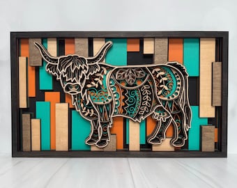May include: A layered wooden art piece featuring a Highland cow silhouette. The cow is intricately detailed with teal and orange accents. The background has a geometric design with orange, teal, and natural wood tones, all within a dark frame.