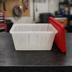 May include: A rectangular, semi-transparent storage bin with a textured, layered surface finish. The bin features a wide, open top and a separate, solid red plastic lid that is positioned leaning against the side of the container.