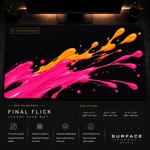 May include: A black rectangular desk mat featuring a vibrant paint splash design in bright orange and hot pink. The top left corner displays the text PAINT SPLASH SERIES. The mat has stitched edges and a smooth surface with a non-slip rubber base.