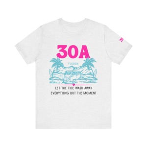 May include: A light grey t-shirt with a graphic design. The design features the text "30A FLORIDA" in pink, with a beach scene in blue. The text "LET THE TIDE WASH AWAY EVERYTHING BUT THE MOMENT" is below the graphic.