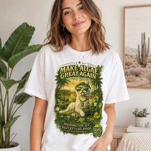 May include: A white t-shirt featuring a graphic print of a cat wearing sunglasses and a gold chain, holding a green drink. The text reads "MAKE ALGAE GREAT AGAIN" and "WASHINGTON D.C. REFLECTING POOL THE NEW GREEN DEAL" with a QR code in the corner.