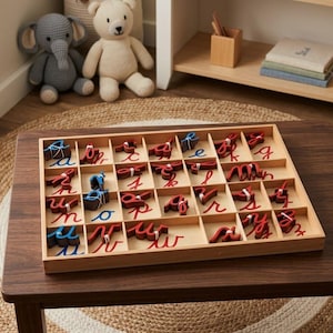 May include: Wooden tray with individual compartments filled with red and blue cursive letters. The letters are arranged in a grid, perfect for educational activities. A teddy bear and elephant toy are in the background.