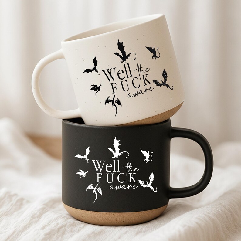 May include: Two ceramic mugs, one cream and one black, stacked on top of each other. Both mugs feature the phrase "Well the FUCK aware" in a stylized font, surrounded by white dragon illustrations. The mugs have a brown base and black handles.