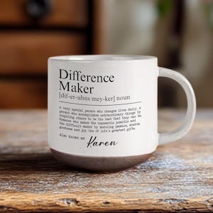 Può includere: Una tazza in ceramica bianca con finitura maculata e base marrone scuro. La parte anteriore presenta un testo nero che definisce il termine Difference Maker, inclusa una pronuncia fonetica e un paragrafo descrittivo, seguito dal nome Karen.