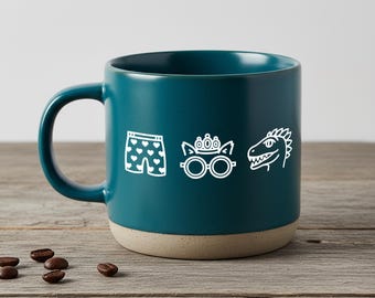 May include: A teal ceramic mug with a matte finish and a light beige base. The front features three white line-art icons: a pair of heart-patterned boxer shorts, a pair of round glasses topped with a small crown, and a dinosaur head profile.