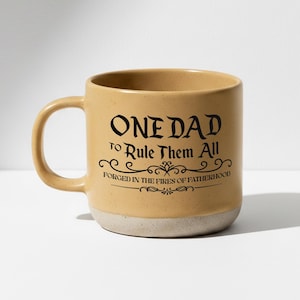 May include: Tan ceramic mug with the text "ONE DAD to Rule Them All FORGED IN THE FIRES OF FATHERHOOD" in black. The mug has a light beige base and a curved handle.